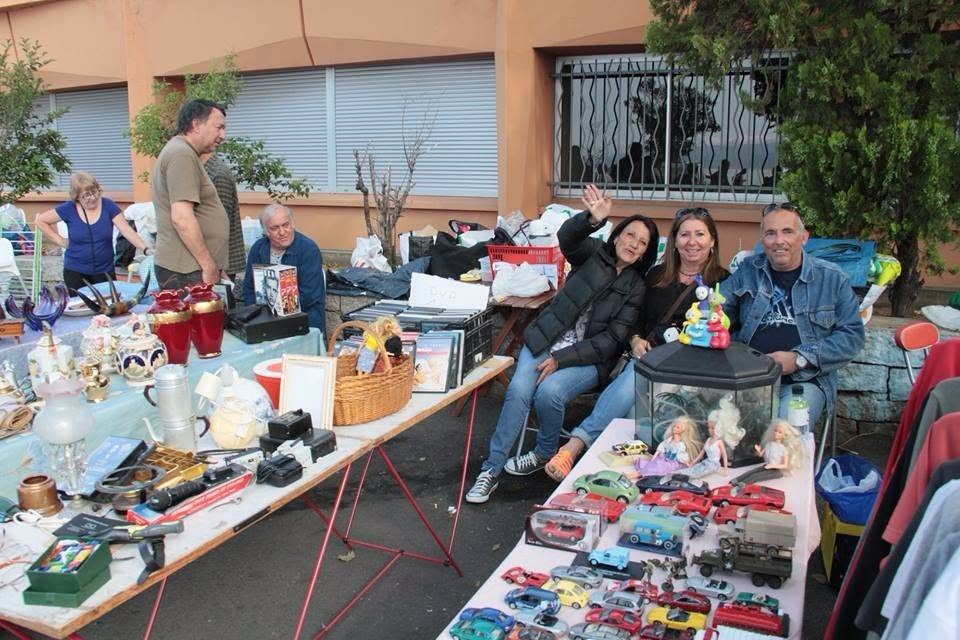 Le stand aux puces : tables couvertes de bibelots, petites voitures, poupées et DVD, avec la famille souriante derrière